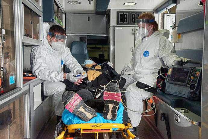 First responders in protective gear, face masks and shields taking a peron's vitals in the back of an ambulance
